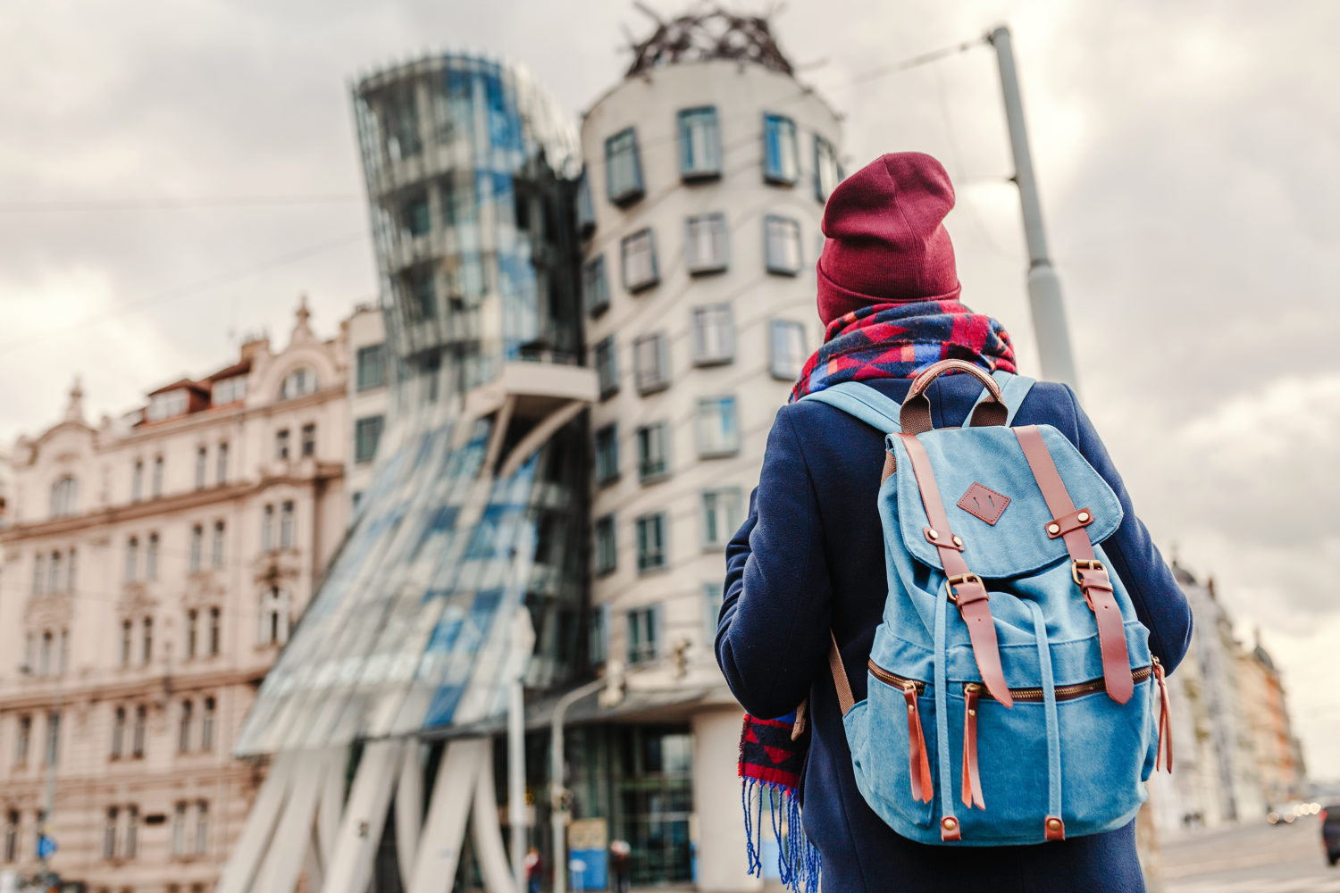 Young hitchhiker in a red hat with a blue backpack standing on a side of a road in Prague