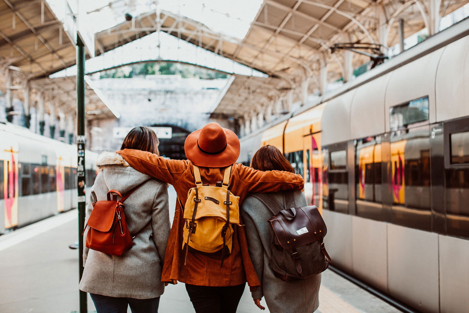 Three women with backpacks walking on a train platform with trains on both sides